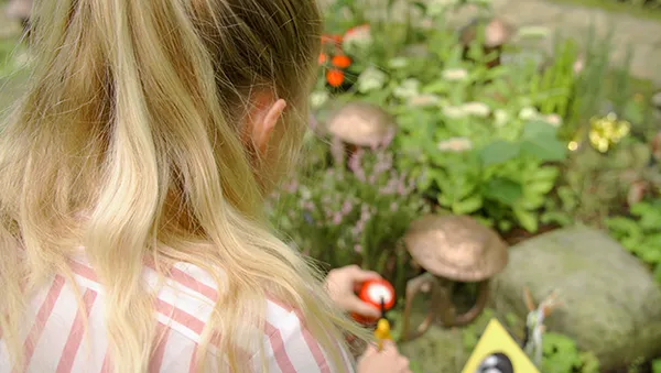 Eine Person mit langen blonden Haaren pflegt einen Garten, umgeben von grünen Pflanzen und Pilzen, und hält dabei einen kleinen rot-schwarzen Gegenstand in der Hand.