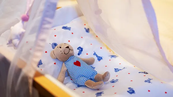 Ein Teddybär in einem blau gestreiften Overall mit einem roten Herzen liegt auf einem Babybett mit einem weiß-blauen Laken mit Tiermuster unter einem durchsichtigen Baldachin.