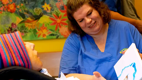 A caregiver in blue scrubs smiles at a person in a colorful hat, holding a painting. A vibrant floral painting is in the background.