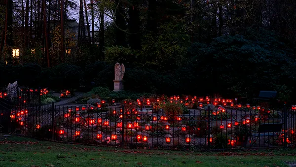 A garden at dusk with numerous red lanterns illuminating pathways, surrounded by trees and a statue of an angel in the background.