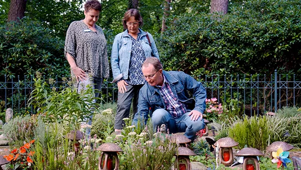 Drei Personen in einem Garten, die Pflanzen beobachten. Ein Mann kniet, während zwei Frauen in der Nähe stehen. Laternen und Grünpflanzen umgeben sie.