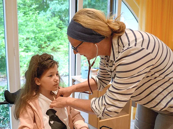 A healthcare professional uses a stethoscope to examine a young girl sitting indoors near a window with greenery outside.
