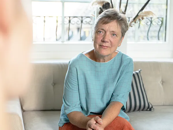Eine ältere Person mit kurzen Haaren sitzt auf einem Sofa, trägt ein hellblaues Oberteil und eine orangefarbene Hose und schaut aufmerksam zu. Im Hintergrund befindet sich ein Fenster.