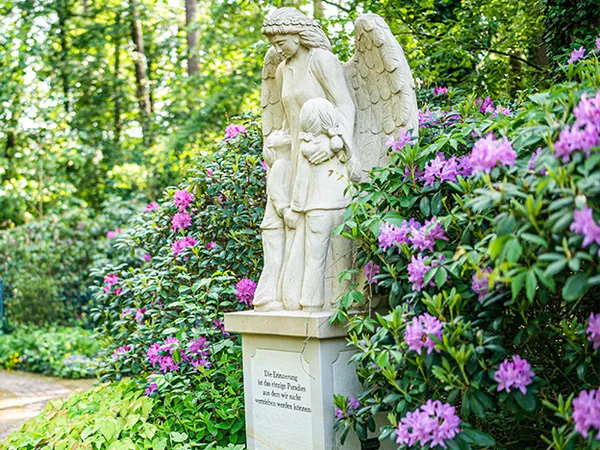 Engelstatue mit Kind, umgeben von leuchtend rosa Blumen und üppigem Grün in einer friedlichen Gartenlandschaft.