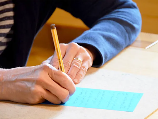 Person, die mit einem Bleistift auf blauem Papier schreibt, einen blauen Pullover und ein gestreiftes Hemd trägt und an einem Holztisch sitzt.