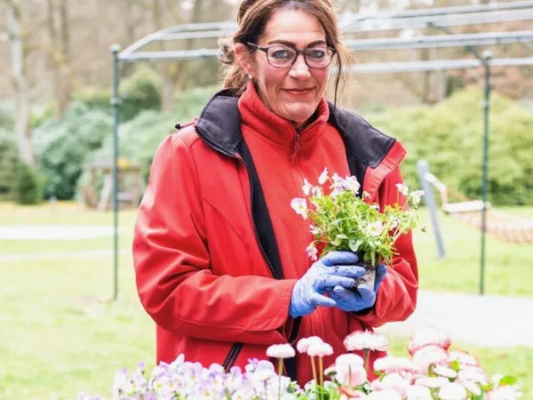 Eine Frau in einer roten Jacke und blauen Handschuhen hält eine kleine Blumenpflanze in der Hand, umgeben von verschiedenen Blumen in einem Garten im Freien.