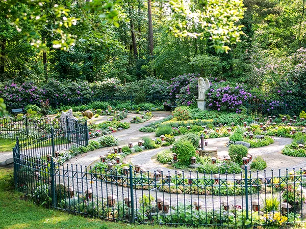 Üppiger Garten mit gewundenen Wegen, leuchtenden Blumen, einer Statue und einem schwarzen Zaun, umgeben von dichtem Grün und blühenden Sträuchern.