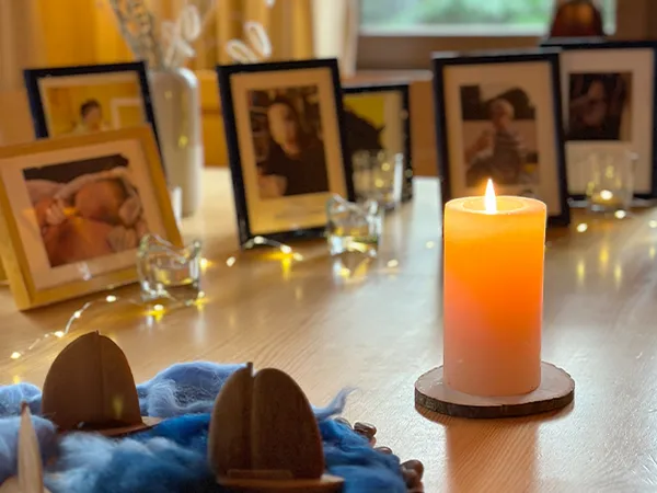 Eine brennende Kerze auf einem Holztisch, umgeben von gerahmten Fotos, Dekoartikeln und sanfter Beleuchtung, schafft eine warme, gemütliche Atmosphäre.