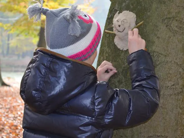 Child in a winter jacket and owl hat creates a clay face on a tree trunk in a forest with autumn leaves.