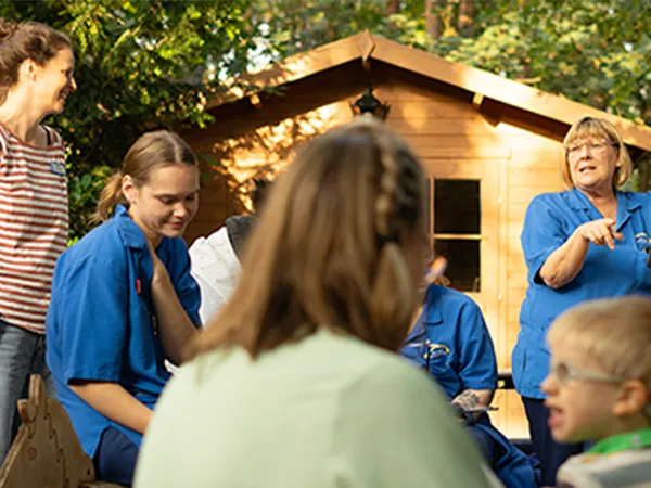 A group of people, including healthcare workers in blue uniforms, are gathered outdoors near a wooden shed, engaging in conversation.