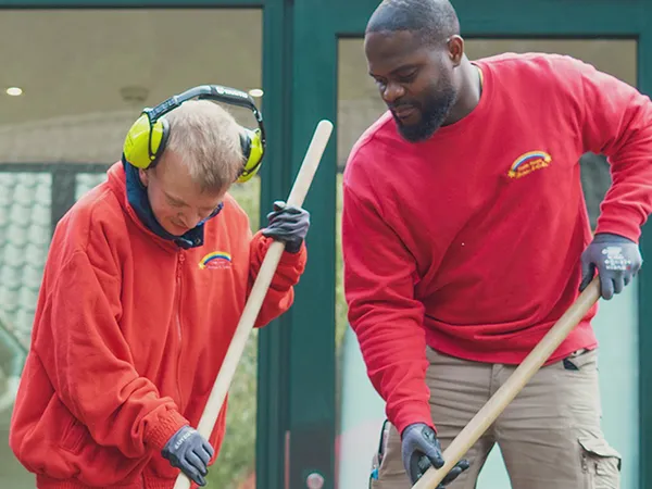 Two people in red shirts raking outdoors, one wearing ear protection. They appear focused on their task in front of a building.