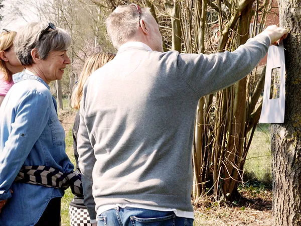 A group of people outdoors, one man holding a paper against a tree, others observing closely. Trees and grass in the background.