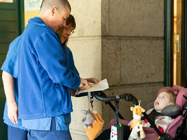 Zwei Personen in blauen Uniformen interagieren mit einem Kind in einem Kinderwagen, an dem Tierfiguren befestigt sind, in der Nähe einer Steinmauer.
