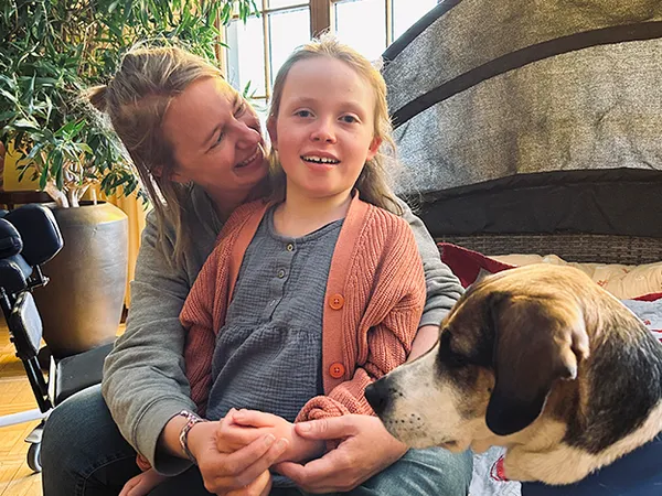 Eine Frau und ein junges Mädchen sitzen zusammen in einem Raum und lächeln herzlich. Ein Hund sitzt in der Nähe und trägt zur gemütlichen Atmosphäre bei.