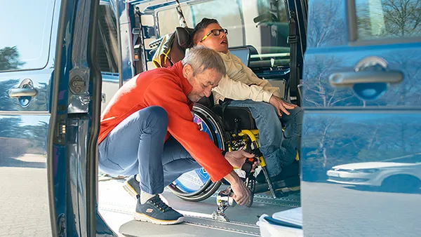 Eine Person hilft einem jungen Rollstuhlfahrer in einem Fahrzeug und passt die Befestigung des Rollstuhls an.