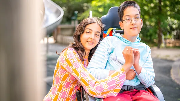 A smiling girl embraces a boy in a wheelchair, both looking at the camera. The background features greenery and a slide.