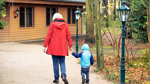 An elderly woman wearing a red jacket runs toward a log cabin, holding a young boy wearing a winter jacket by the hand.