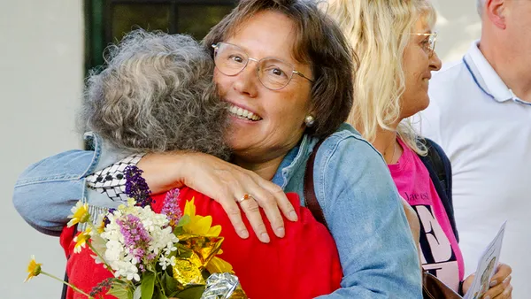 Two women warmly embrace; one holds a bouquet of flowers. A third person stands nearby, smiling.