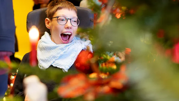 A person with glasses and a scarf smiles joyfully near a decorated Christmas tree with a lit candle in the foreground.