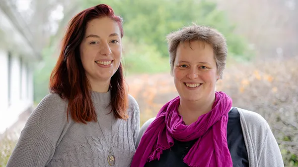 Zwei Personen lächeln im Freien, eine mit roten Haaren und grauem Pullover, die andere mit kurzen Haaren, rosa Schal und schwarzem Oberteil.