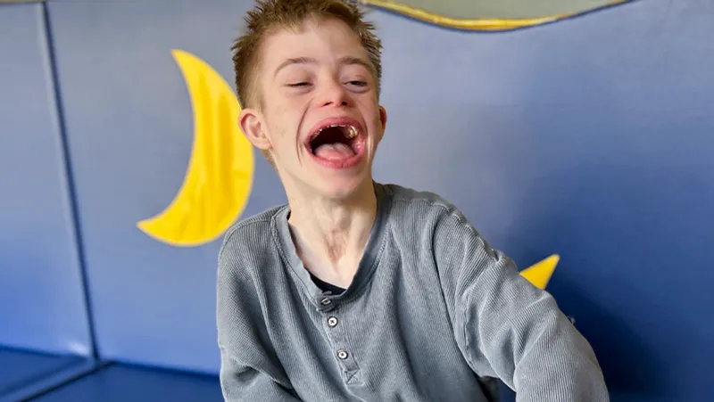 Eine Person mit kurzen Haaren und einem fröhlichen Gesichtsausdruck sitzt vor einer blauen Wand mit gelben Halbmondformen.