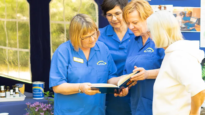 Vier Frauen in blauen Uniformen diskutieren bei einer Veranstaltung in einem Innenraum über ein Buch. Im Hintergrund sind verschiedene Gegenstände und eine Ausstellungstafel zu sehen.