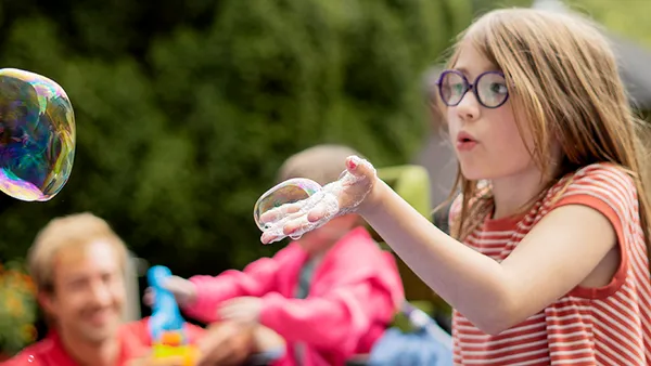 Ein junges Mädchen mit Brille und gestreiftem Shirt bläst draußen Seifenblasen, im Hintergrund sind verschwommene Menschen und Grünpflanzen zu sehen.