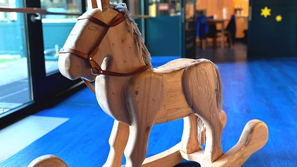 Holzschaukelpferd mit Lederzaumzeug auf blauem Boden, neben einem Fenster mit unscharfem Hintergrund.