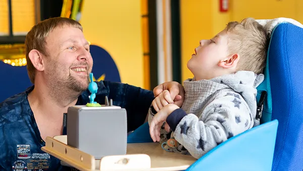 A man smiles warmly at a child in a blue chair, who is wearing a patterned jacket. They are in a brightly lit room with yellow walls.