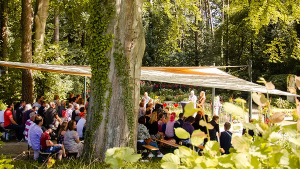 Outdoor gathering under a canopy in a forested area, with people seated on benches, surrounded by lush greenery and dappled sunlight.