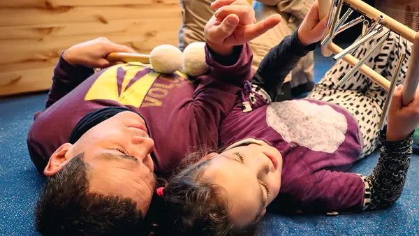 Adult and child lying on the floor, playing musical instruments, wearing casual clothes, and appearing relaxed and joyful.