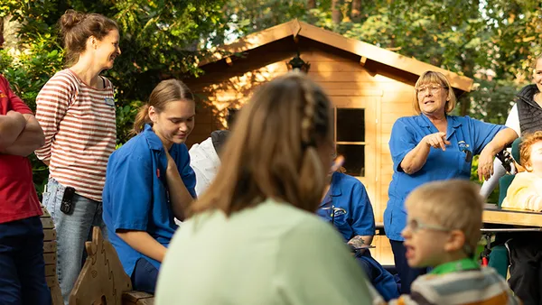 Eine Gruppe von Menschen, darunter eine Frau in einem gestreiften Hemd und andere in blauen Uniformen, versammelt sich im Freien in der Nähe eines Holzschuppens mit Bäumen im Hintergrund.