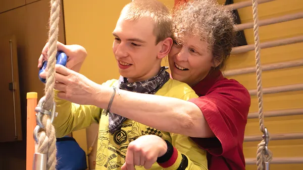 Eine lächelnde Frau hilft einem jungen Mann mit einem Seil in einem hell erleuchteten Raum, beide wirken beschäftigt und glücklich.