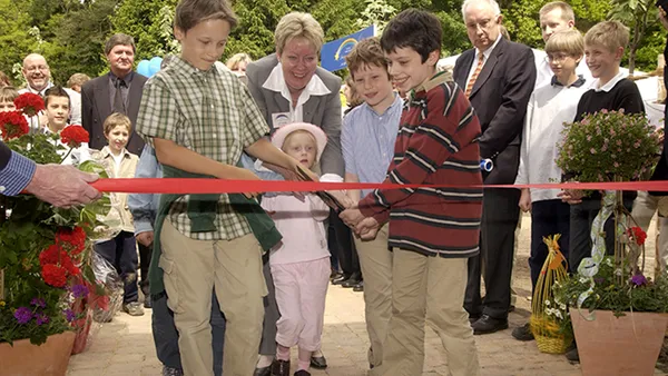Children cut a red ribbon at an outdoor ceremony, surrounded by adults and potted plants, with a crowd watching in the background.