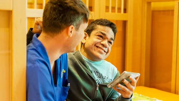 A man in a blue shirt smiles at a boy holding a smartphone, who is also smiling. They are in a warmly lit room.