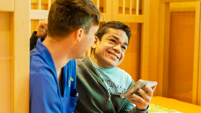 A man in a blue shirt smiles at a boy holding a smartphone, who is also smiling. They are in a warmly lit room.