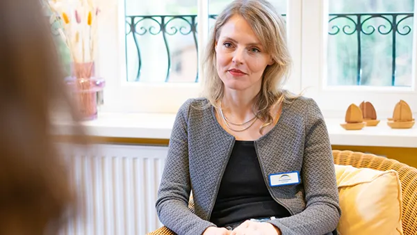 Eine Frau mit blonden Haaren sitzt in einem Korbstuhl, trägt eine graue Jacke und ein Namensschild und befindet sich in einem Raum mit einem Fenster und Dekorationsgegenständen.