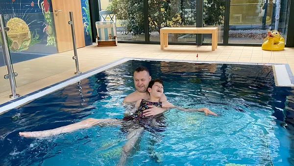 A man supports a child in a swimsuit as they float together in an indoor pool, surrounded by colorful aquatic-themed walls.