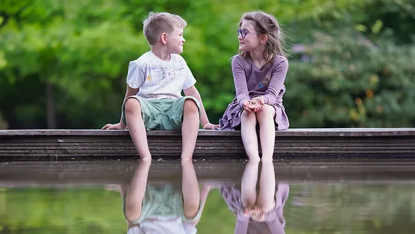 A boy and girl sit on a wooden dock, smiling at each other, with their feet dangling above a reflective pond. Lush greenery surrounds them.