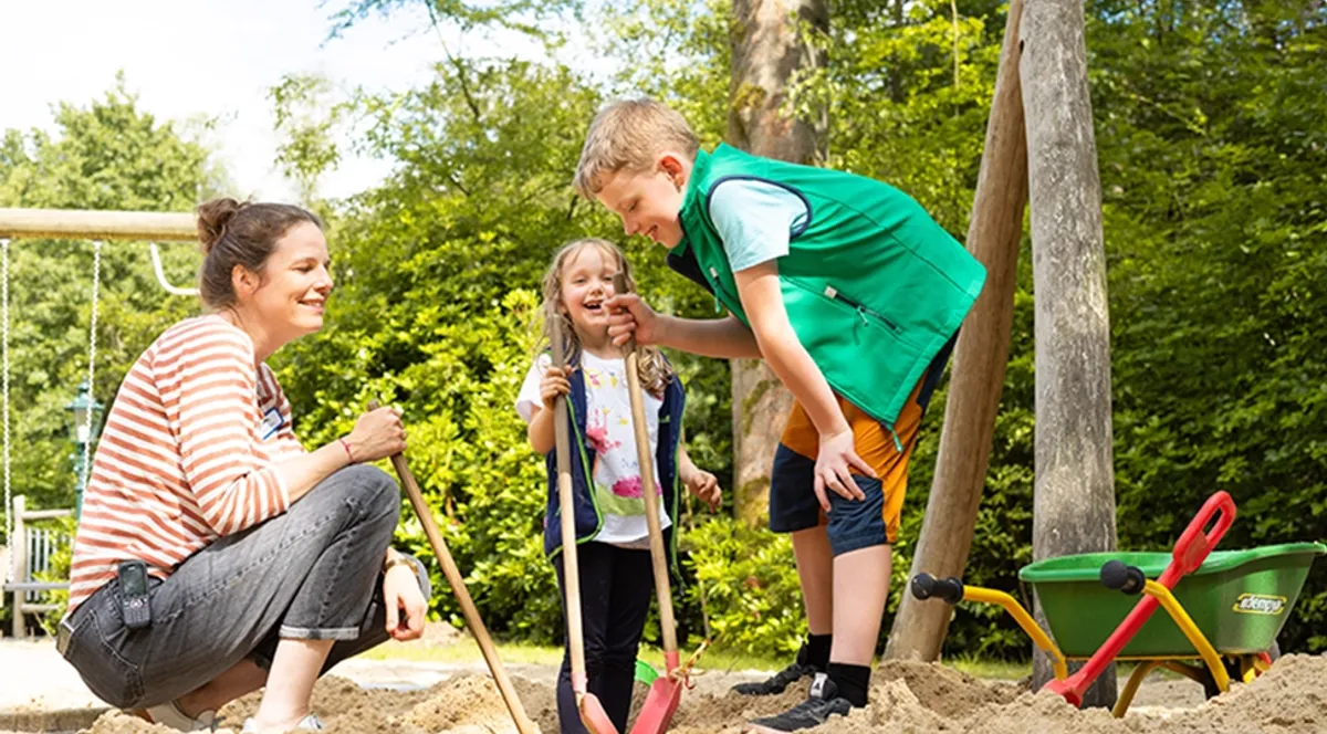 Eine Frau und zwei Kinder spielen mit Schaufeln auf einem sandigen Spielplatz. Im Hintergrund stehen eine Schubkarre und Schaukeln. Der Bereich ist von Bäumen umgeben.