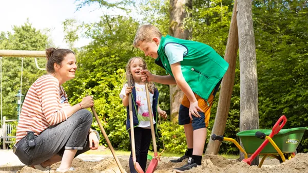Eine Frau und zwei Kinder spielen mit Schaufeln auf einem sandigen Spielplatz. Im Hintergrund stehen eine Schubkarre und Schaukeln. Der Bereich ist von Bäumen umgeben.