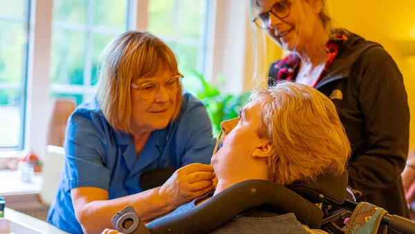 A caregiver in blue assists a person in a wheelchair, while another woman watches, in a warmly lit room with a window in the background.