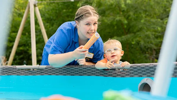 Woman and child leaning on pool edge, woman holding a toy, both smiling. Greenery in the background.