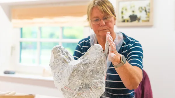A woman wearing glasses and a striped shirt sculpts a large, abstract stone piece in a bright room with a window in the background.
