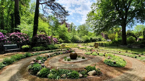 Ein ruhiger Garten mit kreisförmigen Steinwegen, üppigem Grün, blühenden Blumen und einer zentralen kugelförmigen Skulptur unter einem teilweise bewölkten Himmel.