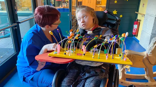 Eine Pflegekraft interagiert mit einem Kind im Rollstuhl. Das Kind beschäftigt sich mit einem bunten Sensorikbrett.