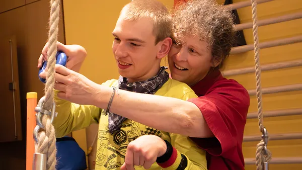 A smiling woman assists a young man with a rope in a brightly lit room, both appearing engaged and happy.