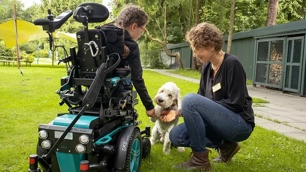 A person in a wheelchair pets a white dog while another person kneels beside them on grass, with trees and a building in the background.